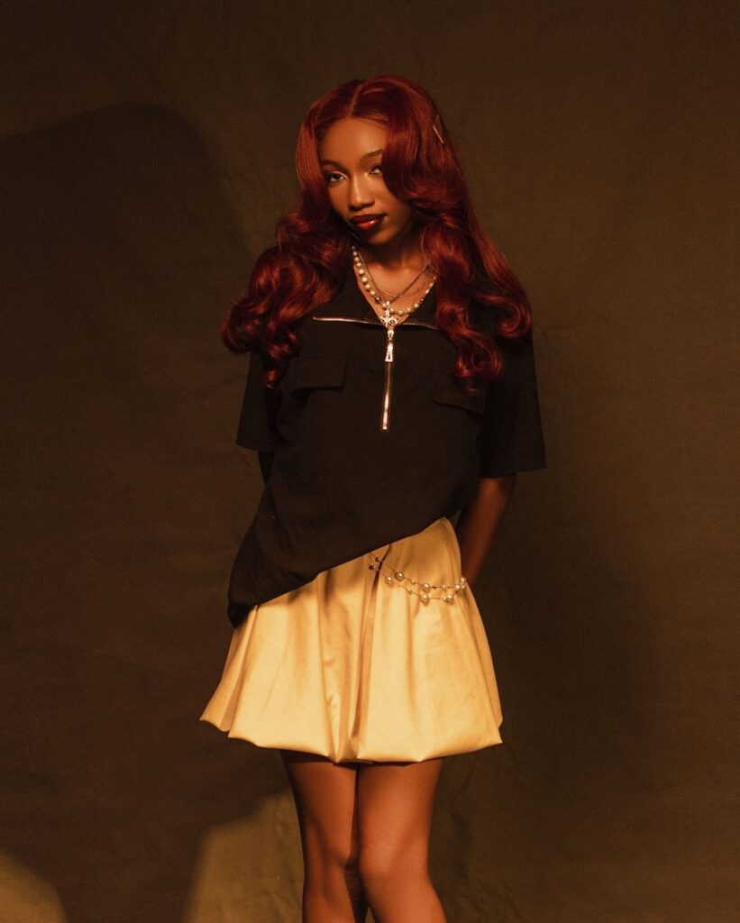 Fewa in a black zip shirt, pearls and a pale bubble skirt, red waves over one shoulder, warm amber studio light