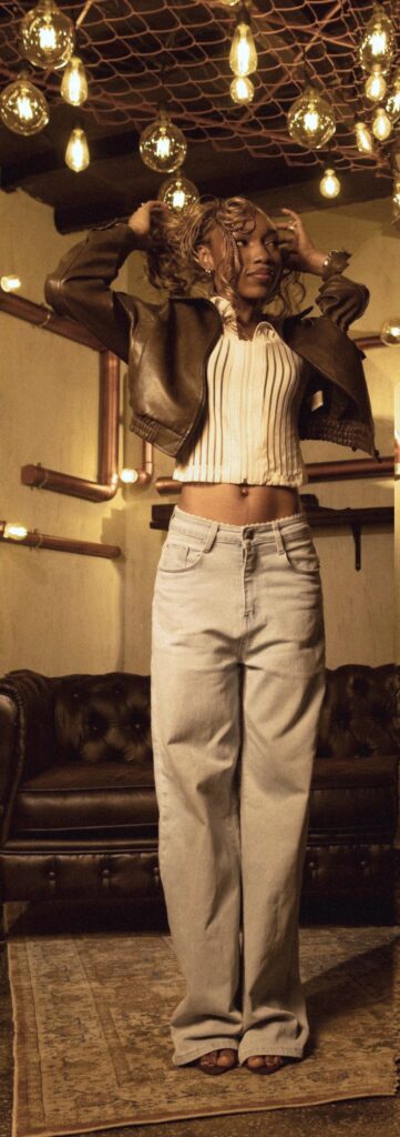 Fewa lifting her curls with both hands, leather jacket open over a cropped striped top