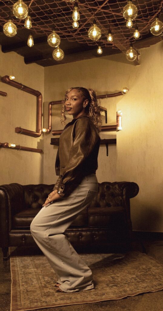 Fewa mid-pose in a leather jacket, knees bent, glancing back over her shoulder