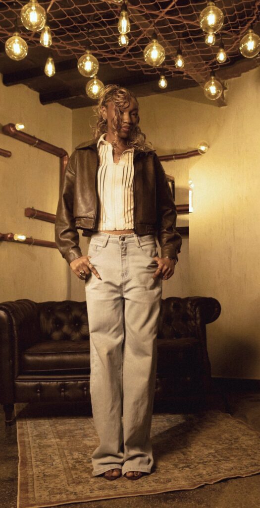 Fewa standing tall in a leather jacket and wide jeans, thumbs hooked in her pockets, looking down at the camera