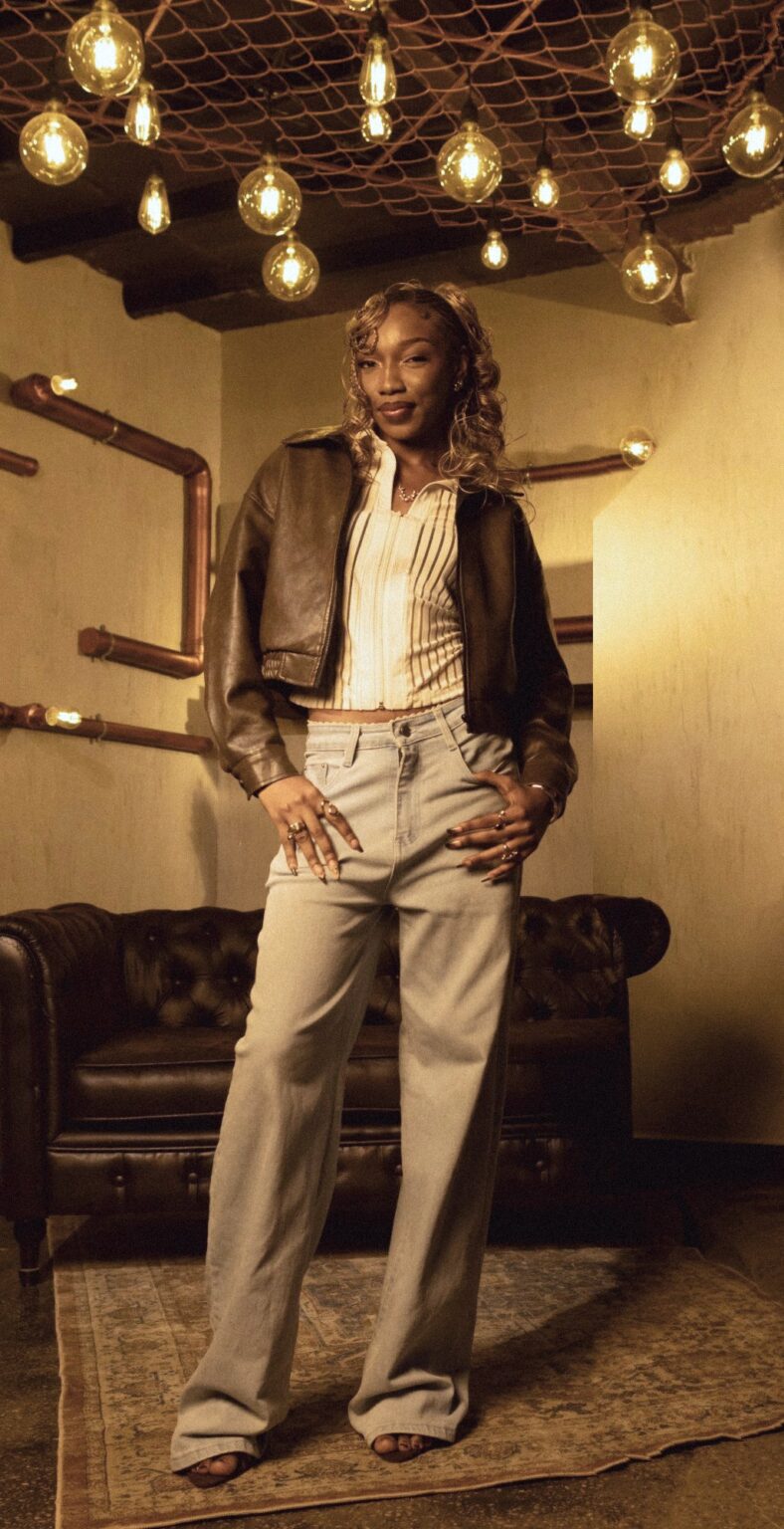 Fewa in a brown leather bomber, striped top and wide light-wash jeans, hands on hips beneath hanging Edison bulbs