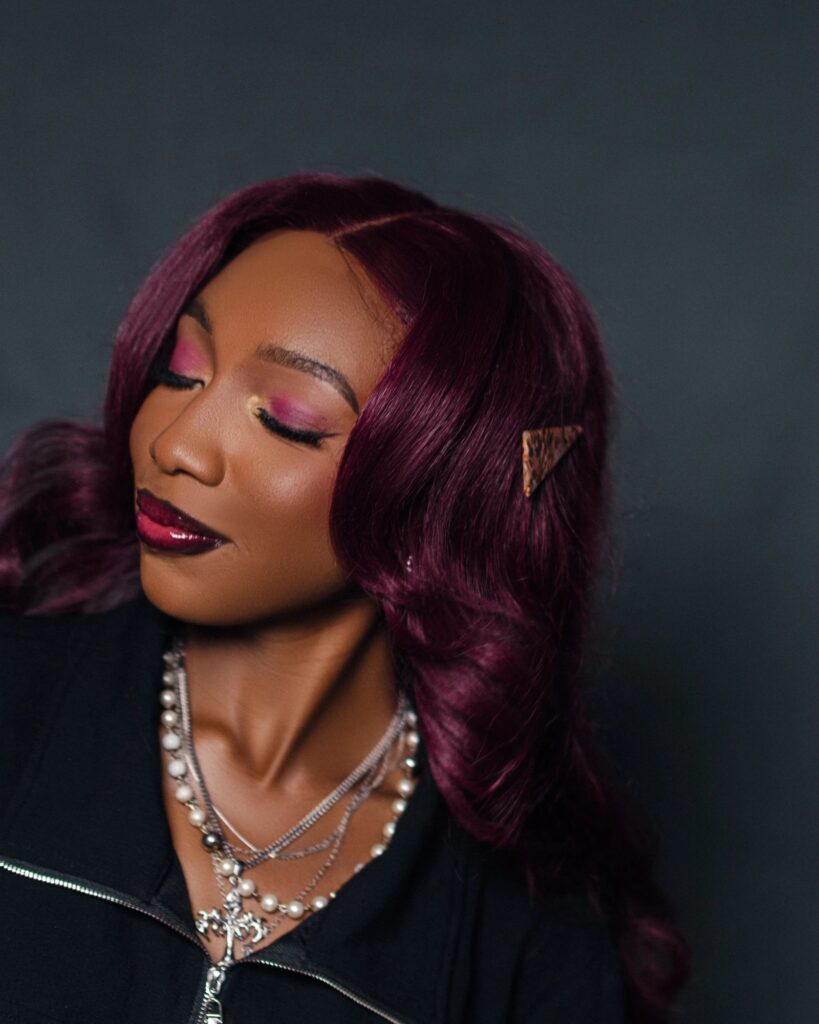 Fewa with plum-coloured hair, eyes closed, pink eyeshadow and layered pearl and silver necklaces