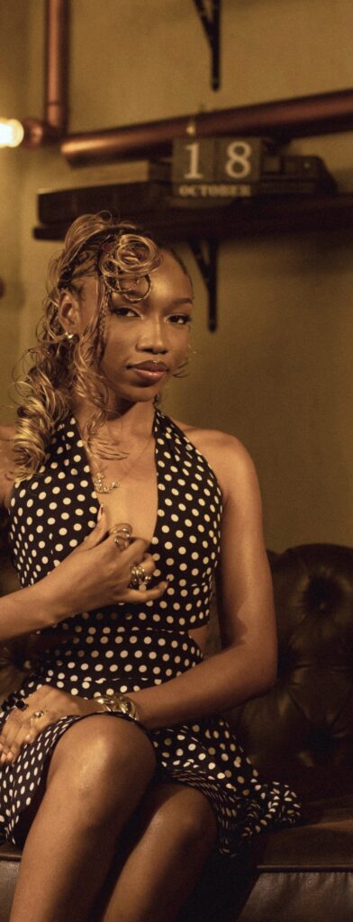 Fewa in a navy polka-dot halter dress on a leather Chesterfield, one hand at her chest, gold rings and cuff