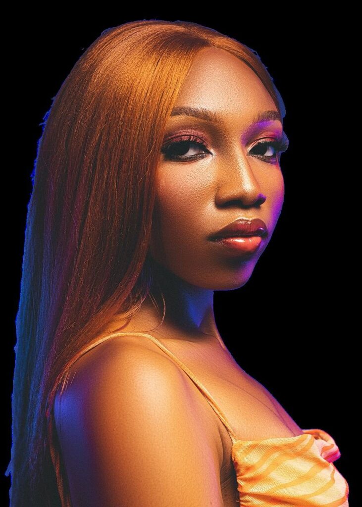 Fewa in a satin orange top with long auburn hair, lit in warm orange against black
